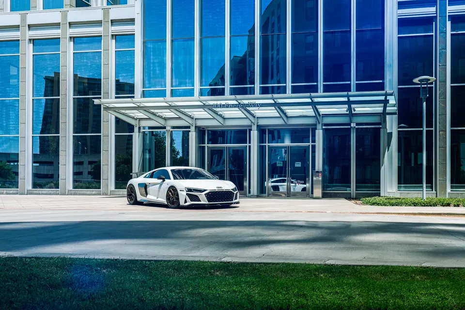 White Audi R8 sports car parked in front of a modern glass office building on a sunny day.