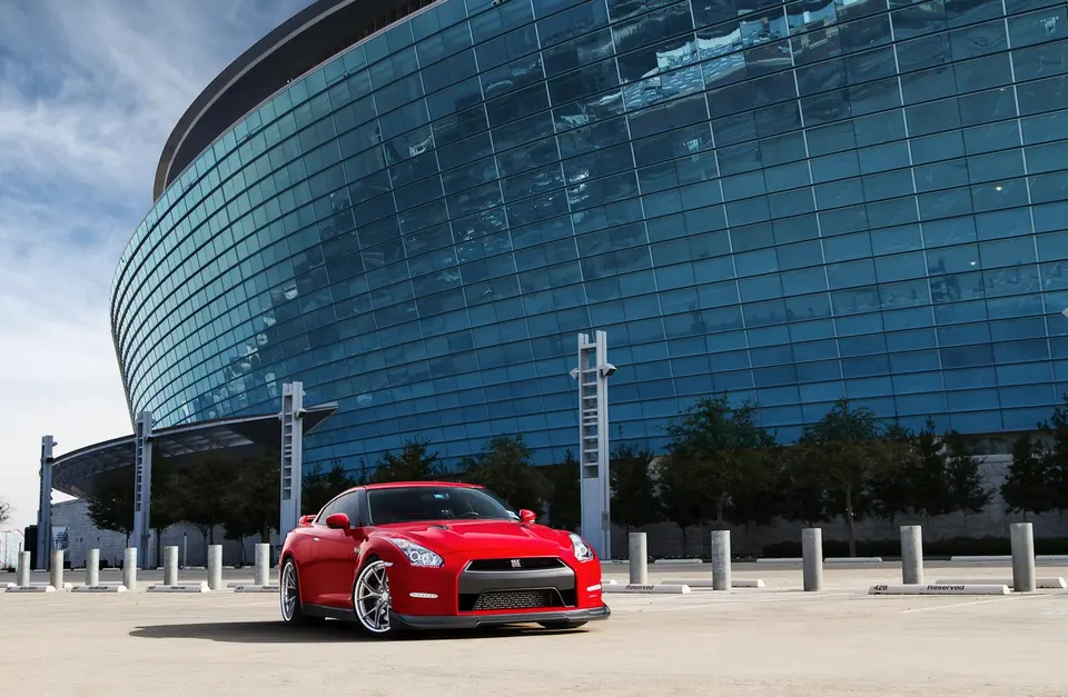Red Nissan GT-R R35 with custom wheels parked in front of a modern glass building on a sunny day.