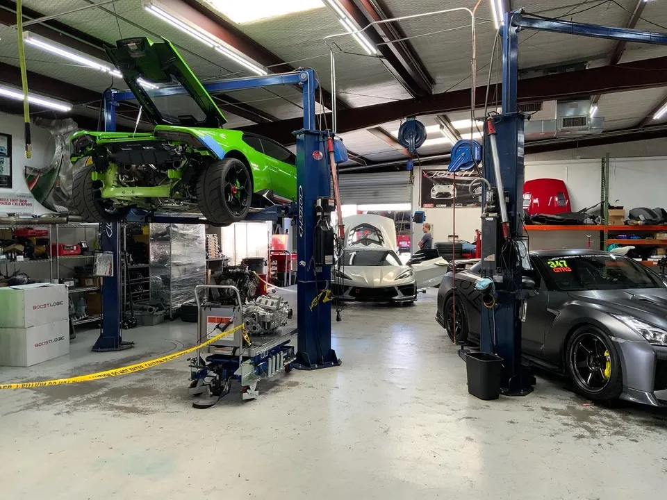 A busy performance tuning shop with a green Lamborghini Huracan on a lift, a grey Nissan GT-R, and a white Corvette C8.