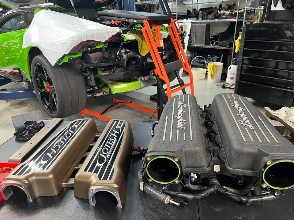 A green Lamborghini Huracan undergoing an engine build in a Jotech Motorsports shop, with stock and aftermarket intake manifolds on display.