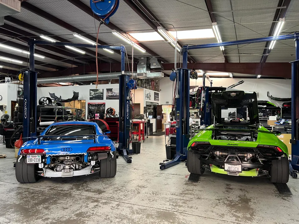 Blue Audi R8 and green Lamborghini Huracan with exposed rear turbo and exhaust systems on lifts in a performance tuning shop.