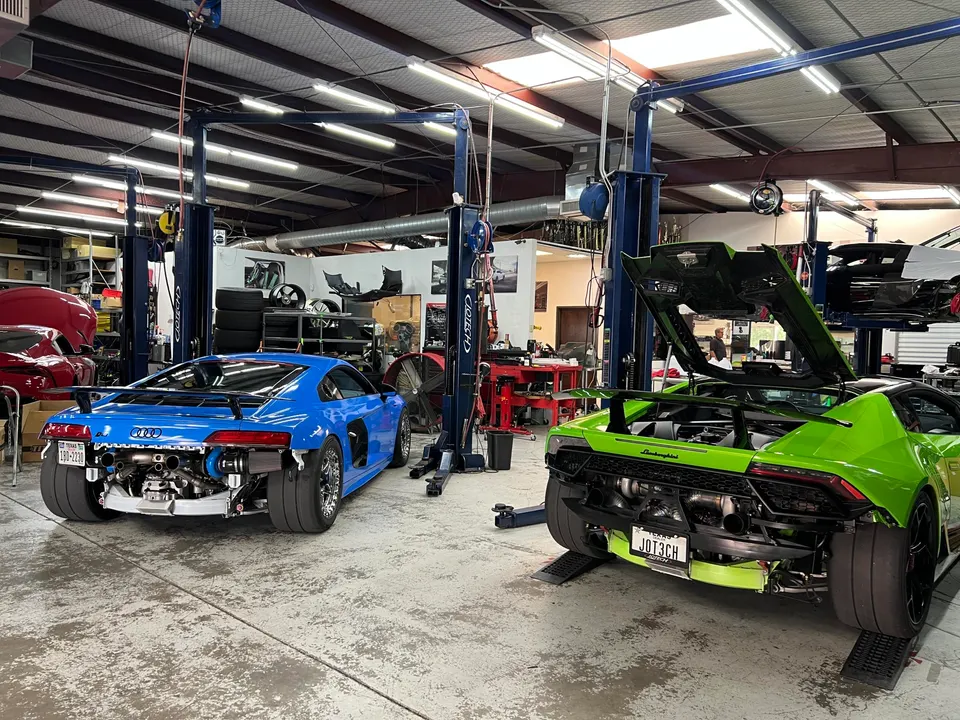 Two modified supercars, a blue Audi R8 and a green Lamborghini Huracan, with exposed twin-turbo setups in a performance shop.