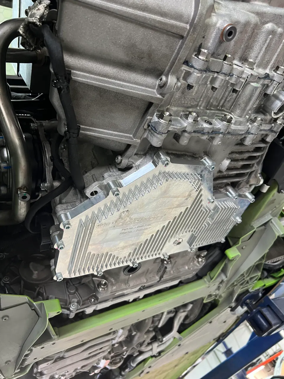 A detailed view of a billet transmission pan installed on the underside of a Lamborghini in a performance shop.
