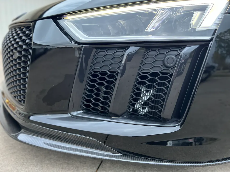 Close-up of a black Audi R8 front bumper with a carbon fiber splitter, honeycomb grille, and visible intercooler.