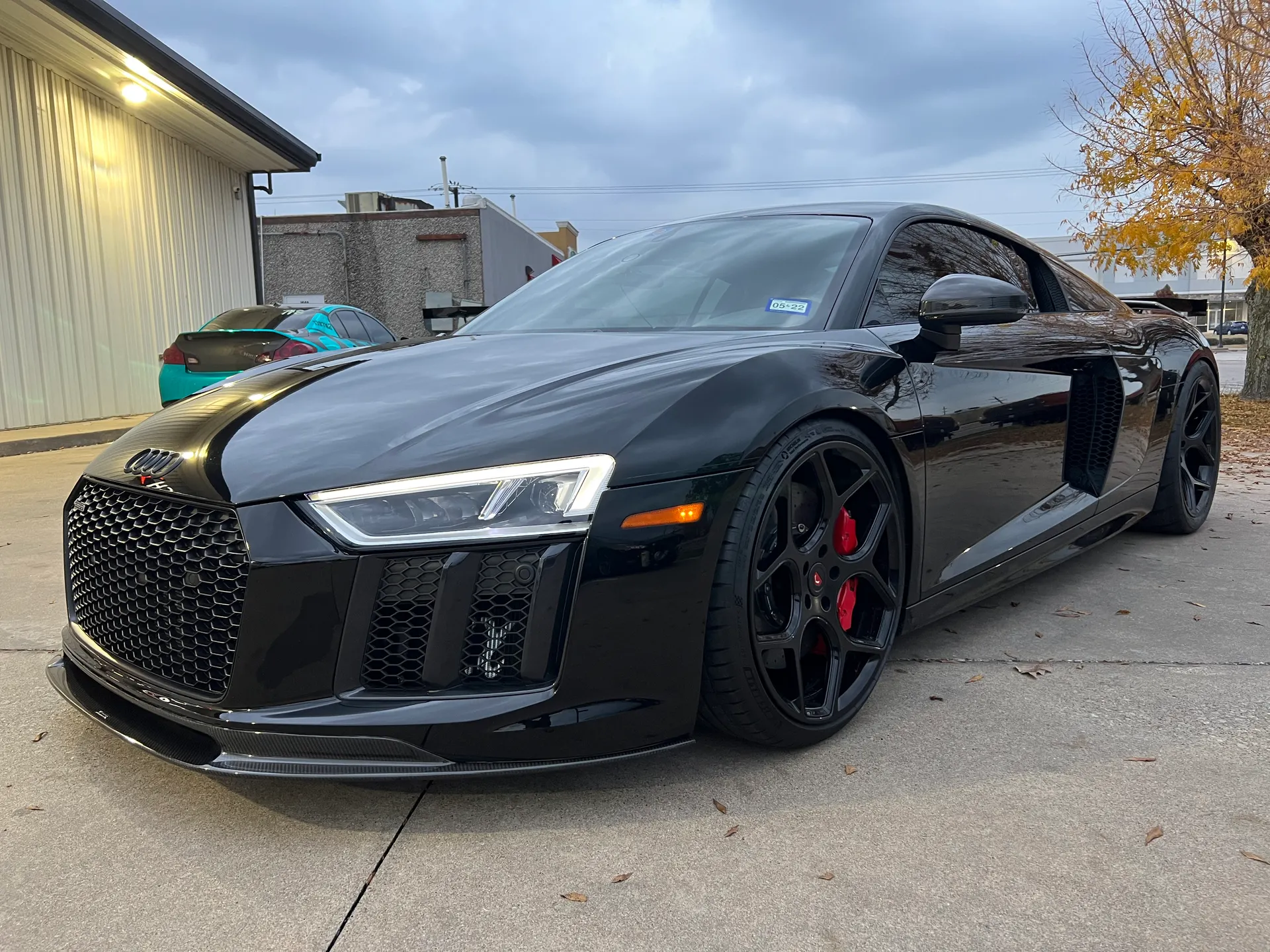 A black Audi R8 with a carbon fiber front splitter and aftermarket wheels parked outside a building.