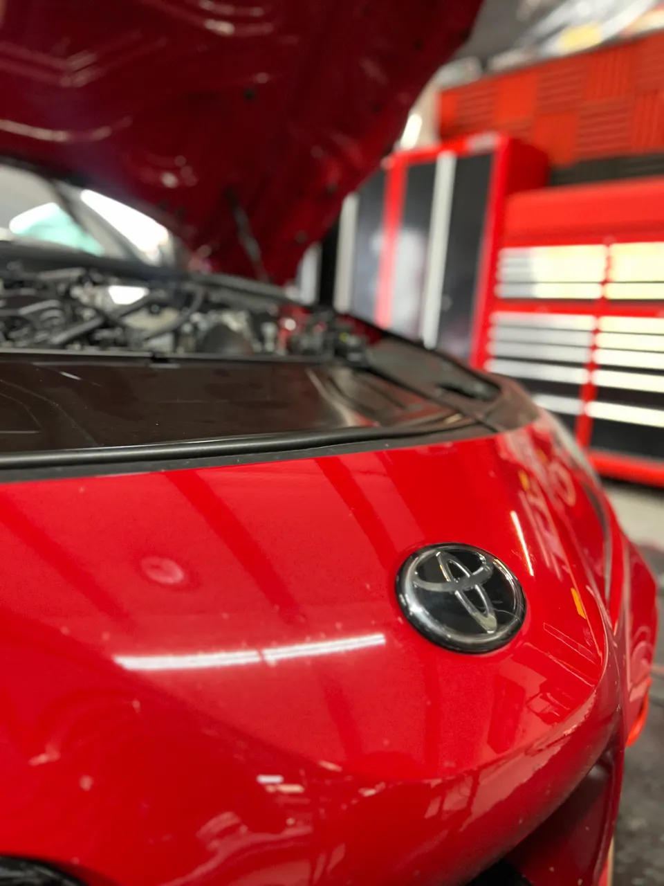 A red Toyota Supra with its hood open in a performance tuning shop, with red toolboxes visible in the background.
