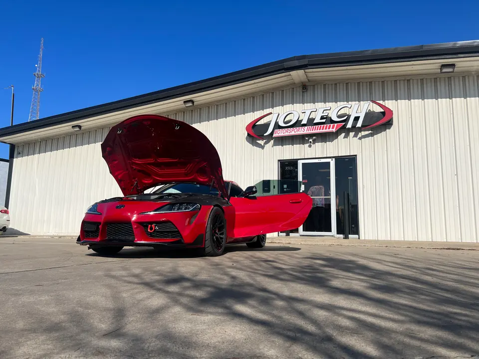 A red Toyota Supra with its hood open parked outside Jotech Motorsports performance tuning shop.