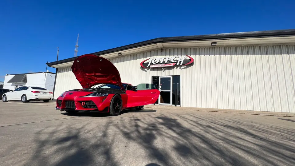 A red Toyota Supra with its hood and driver's door open, parked outside Jotech Motorsports performance tuning shop.