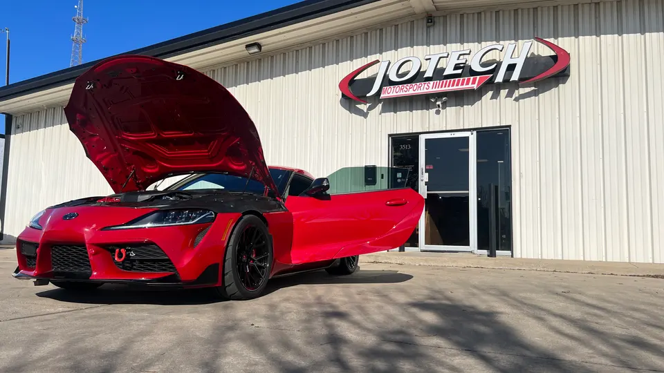 Red Toyota Supra with hood and driver's door open, parked outside Jotech Motorsports performance tuning shop.