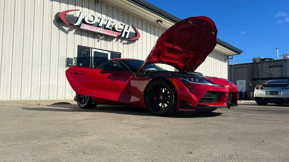Red Toyota Supra with its hood open parked outside Jotech Motorsports, with a silver Nissan GT-R in the background.