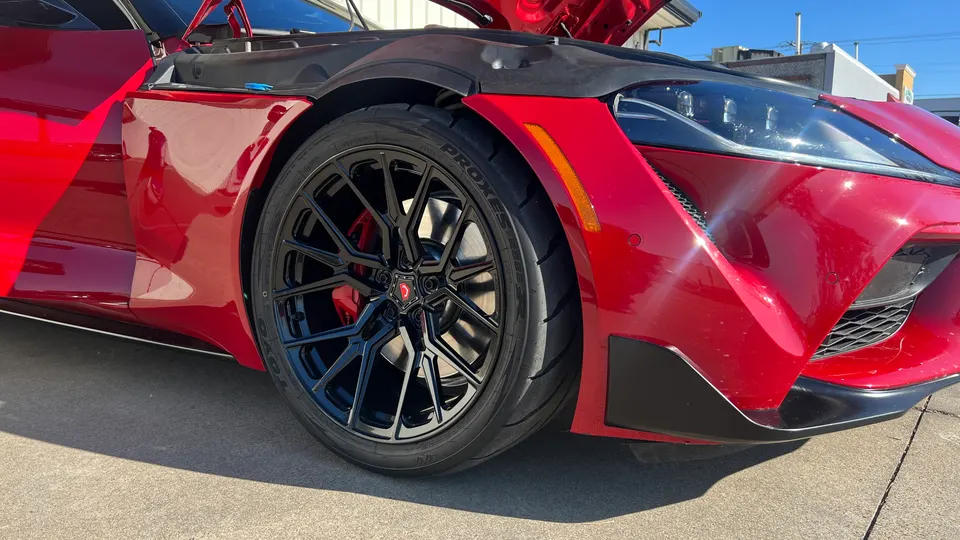 Close-up of a red Toyota Supra's front wheel, featuring black aftermarket wheels, Toyo Proxes tires, and red brake calipers.