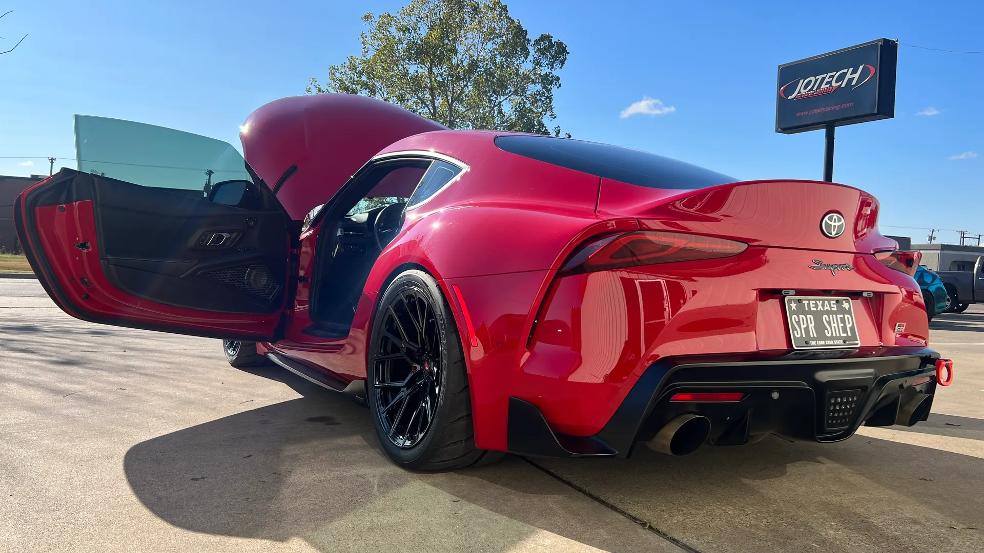 Red Toyota Supra with custom black wheels parked outside Jotech Motorsports with hood and driver's door open.