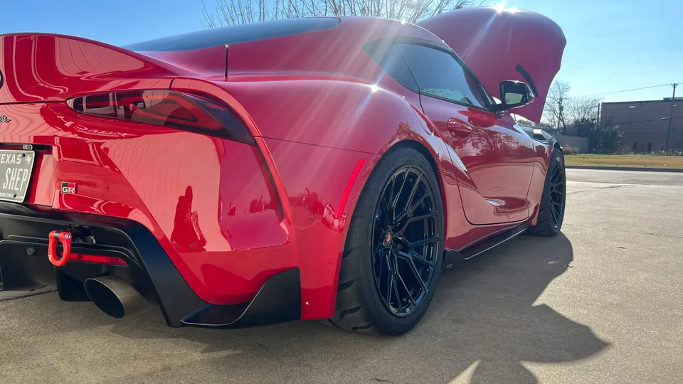 Red Toyota Supra with aftermarket black wheels, a visible exhaust tip, and a red tow hook, parked outdoors with its hood open.