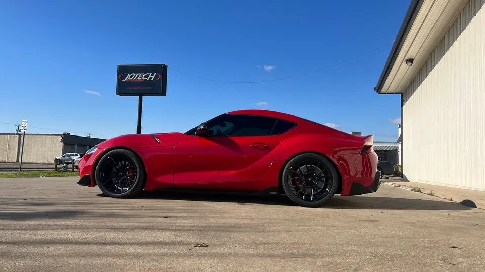 A red Toyota Supra with black aftermarket wheels parked outside the Jotech Motorsports shop on a sunny day.
