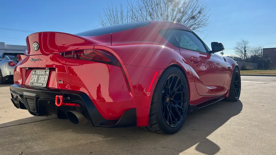 Red Toyota Supra GR with aftermarket black wheels, performance tires, and a red tow hook, parked outdoors on a sunny day.