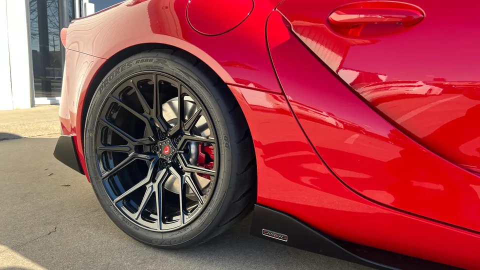 A close-up of the rear wheel and side of a red Toyota Supra with black aftermarket wheels and red brake calipers.