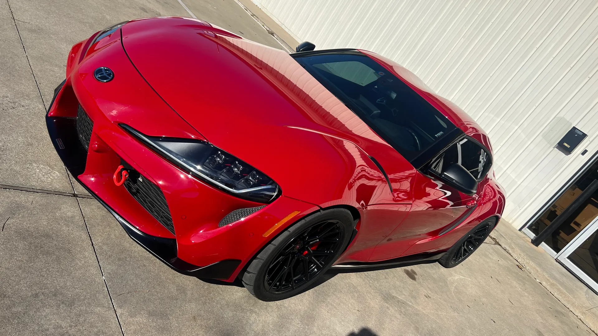 A red Toyota Supra with black aftermarket wheels and a red tow hook parked outside a building on a sunny day.