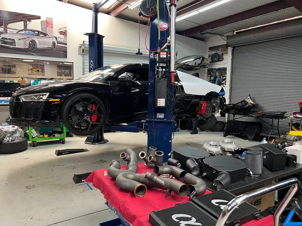 Black Audi R8 on a lift in a performance shop, surrounded by turbochargers, exhaust headers, and other tuning parts.