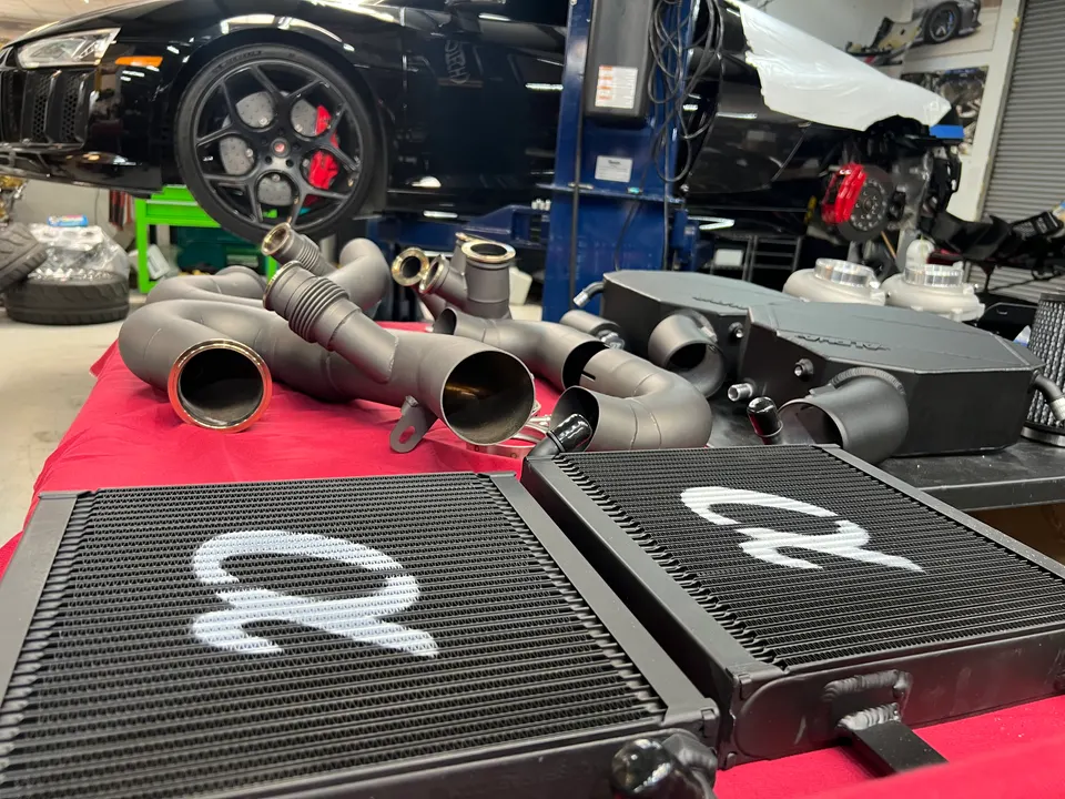 Performance intercoolers and boost pipes on a table in a shop with a black Lamborghini or Audi R8 on a lift.