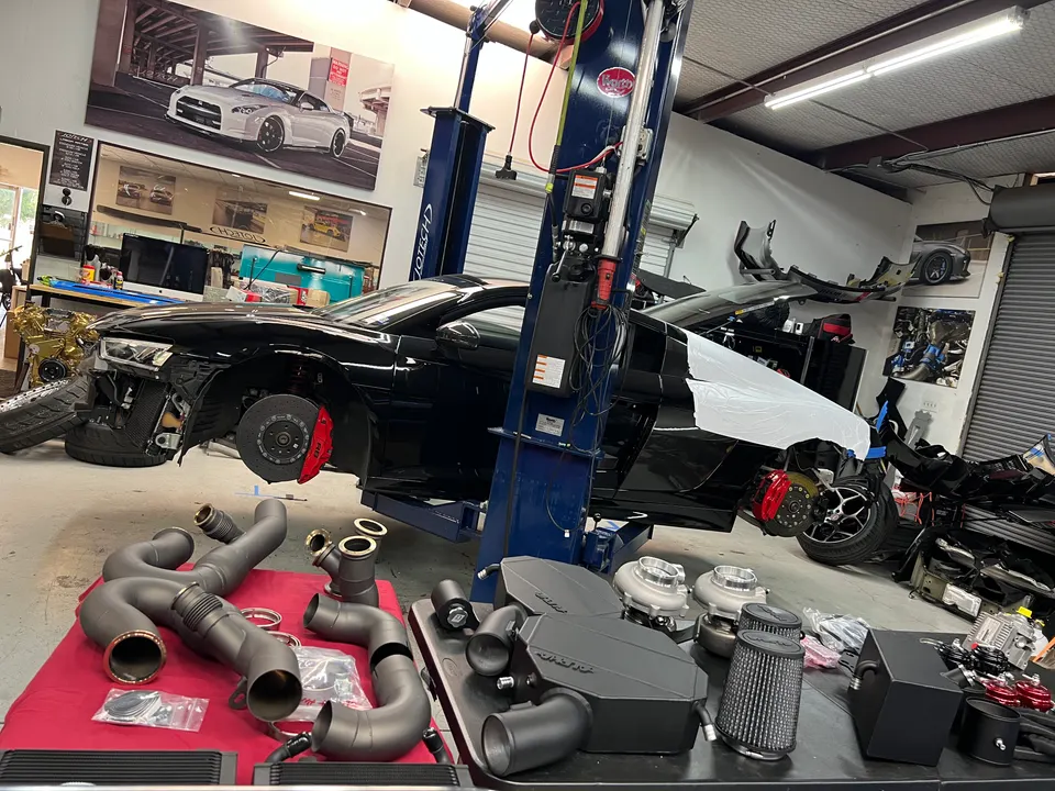 Audi R8 on a lift in a performance tuning shop with turbochargers, intercoolers, and other aftermarket parts laid out.