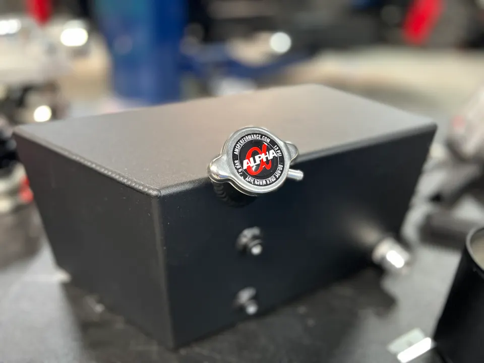 Alpha Performance branded black aluminum coolant overflow tank or catch can on a workbench.