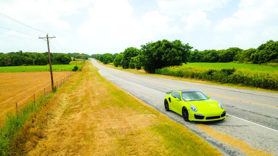 A vibrant lime green Porsche 911 sports car driving on a scenic country road surrounded by fields and trees.