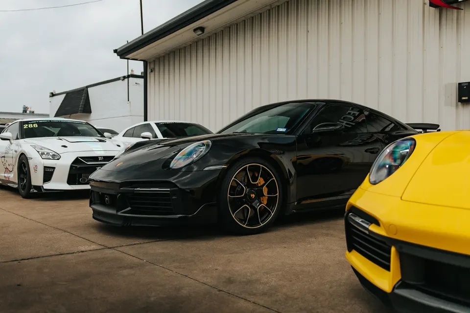 A white Nissan GT-R and a black Porsche 911 parked outside a performance shop, with a yellow Porsche partially visible.