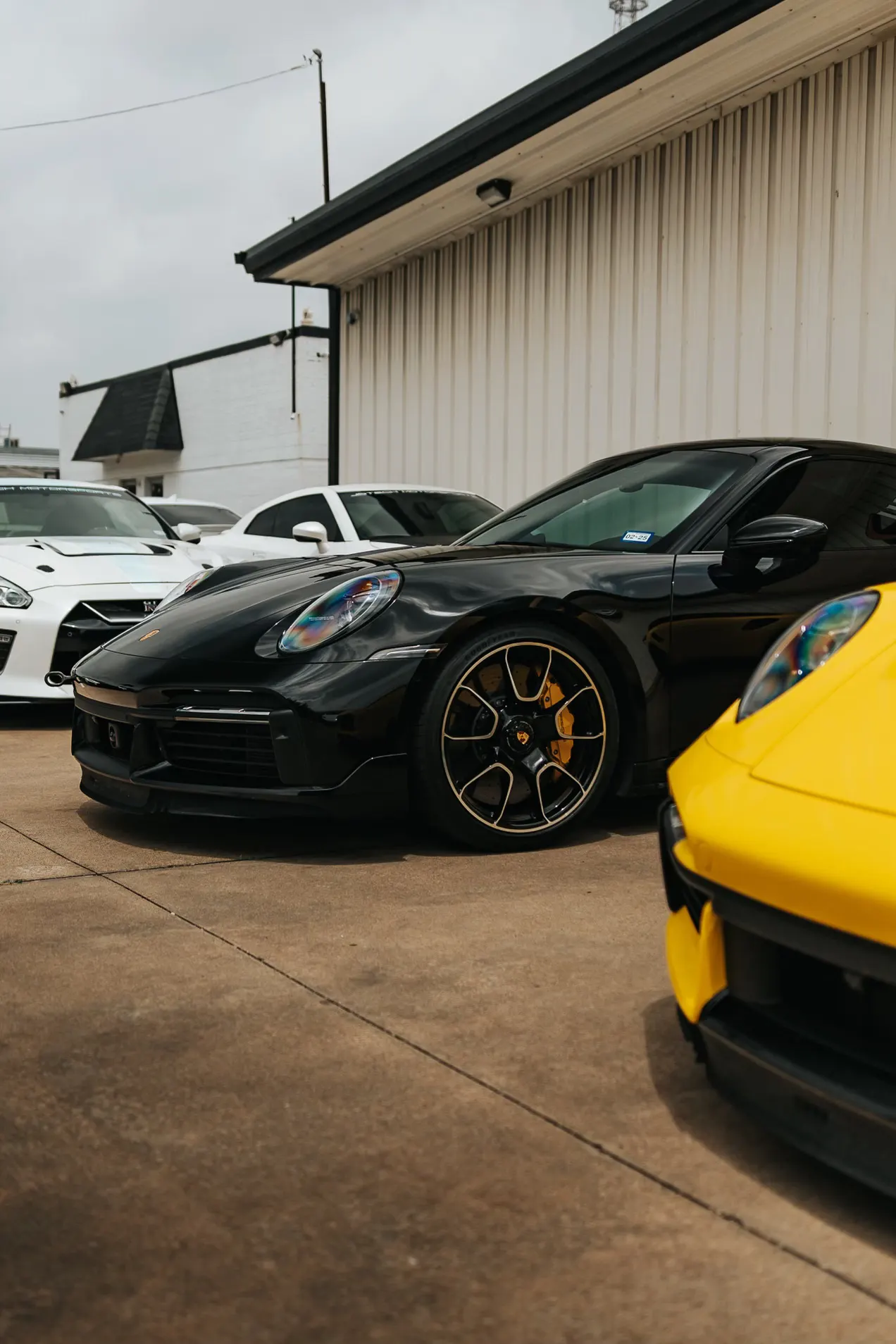 A black Porsche 911, a white Nissan GT-R, and a yellow Porsche parked outside a performance shop.