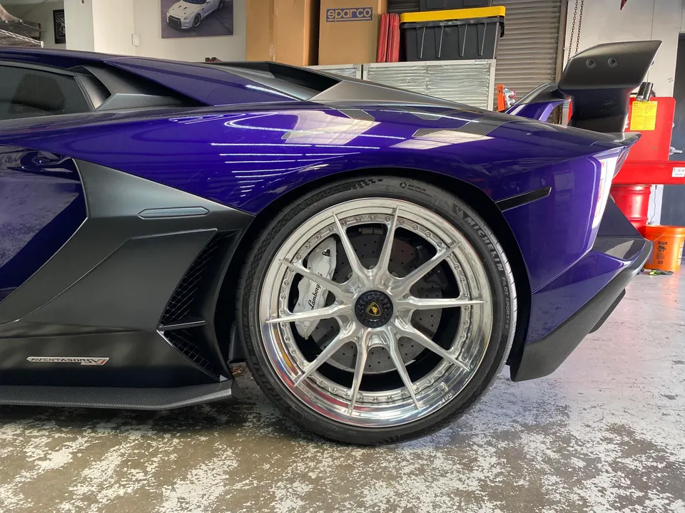 A purple Lamborghini Aventador with custom silver wheels and carbon fiber aero in a shop setting.