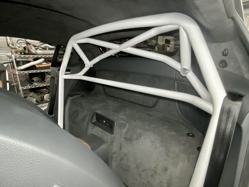 A white roll cage installed in the rear interior of a performance car at a tuning shop, with parts shelves in the background.