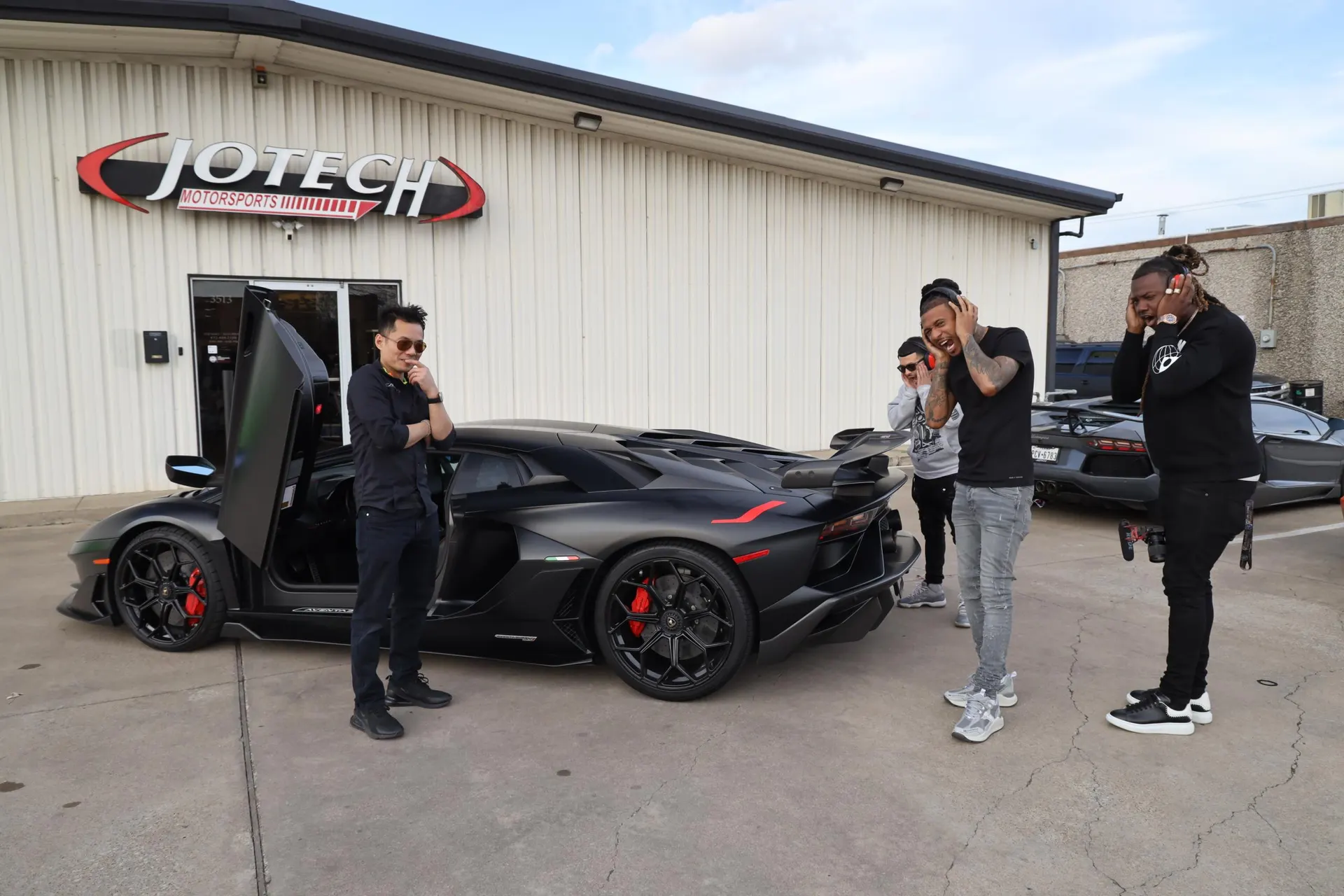Black Lamborghini Aventador with scissor doors open parked outside Jotech Motorsports, surrounded by people.