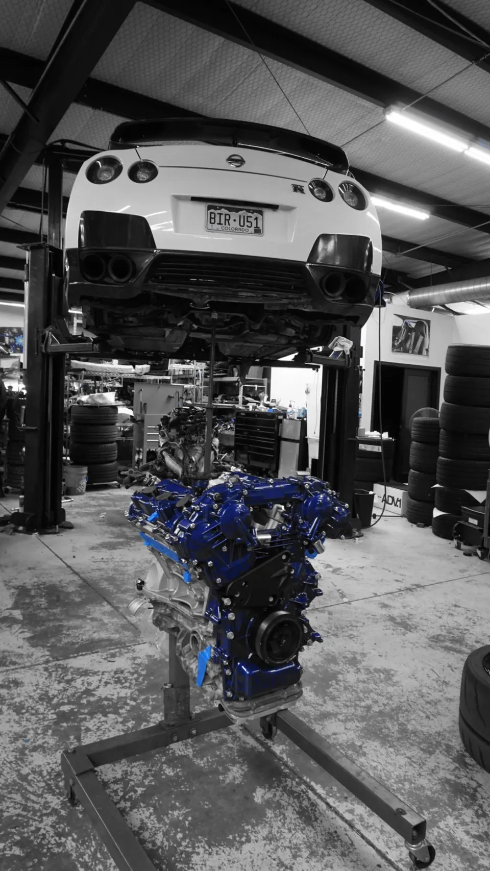 White Nissan GT-R on a lift in a performance shop with a custom blue built engine on a stand below it.