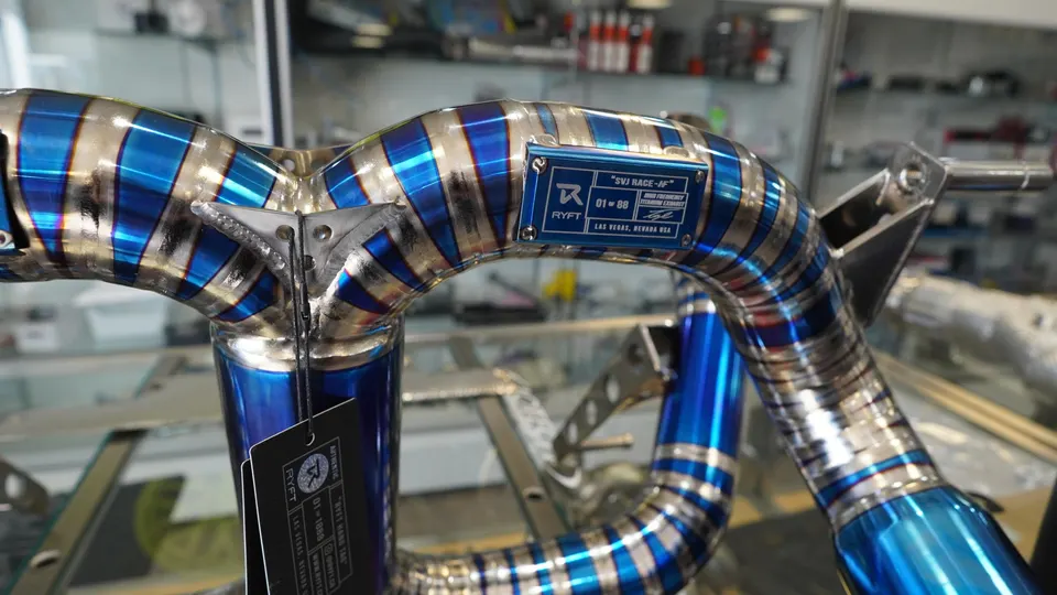 Close-up of a blue and gold titanium exhaust pipe with a Ryft brand tag, displayed in a shop setting.