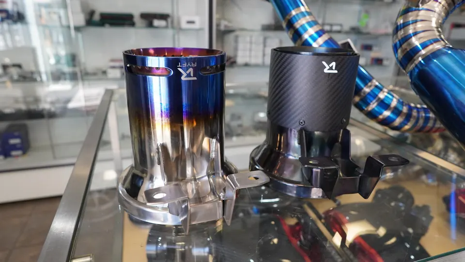 Close-up of two aftermarket exhaust tips, one titanium and one carbon fiber, on a glass display in a performance shop.