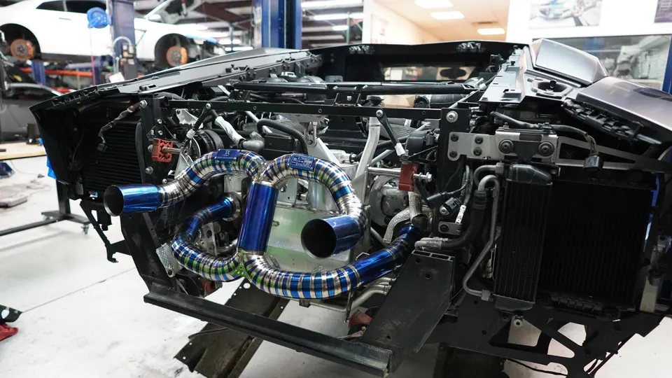 A Lamborghini with its front end removed, showcasing a custom blue titanium exhaust system being installed in a performance shop.