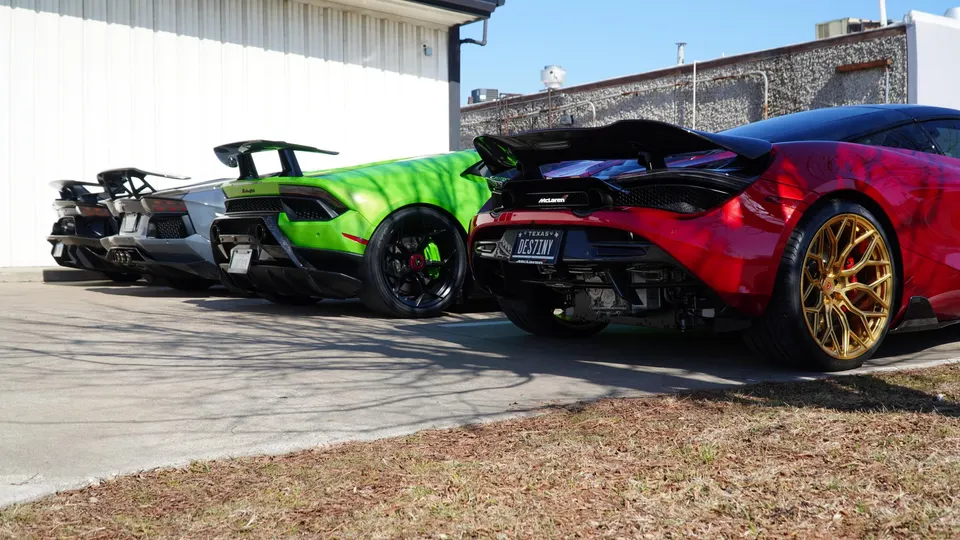 Rear view of multiple supercars, including a green Lamborghini and a red McLaren with gold wheels and large rear wings, parked outdoors.