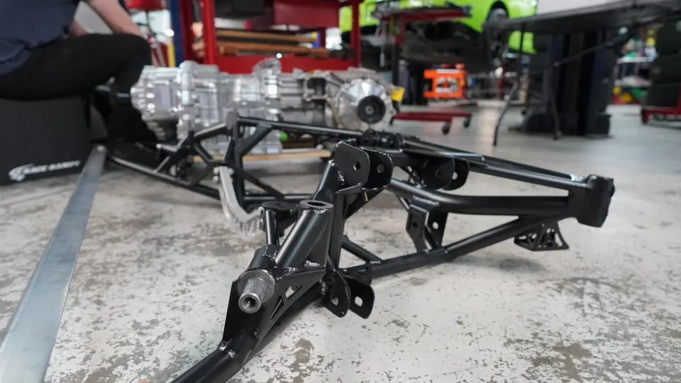 A black car chassis frame and a silver transmission assembly on a concrete floor in a performance tuning shop.