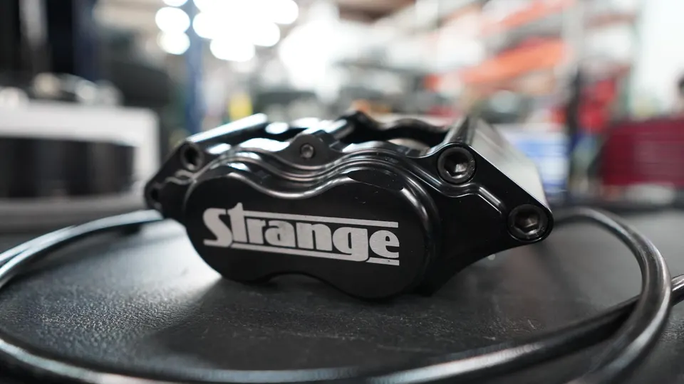 A close-up of a black Strange Engineering performance brake caliper on a workbench in a tuning shop.
