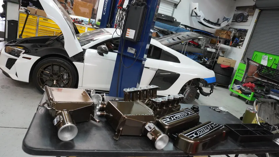 White Audi R8 in a Jotech Motorsports shop with the hood open, surrounded by performance intercoolers and intake manifolds.