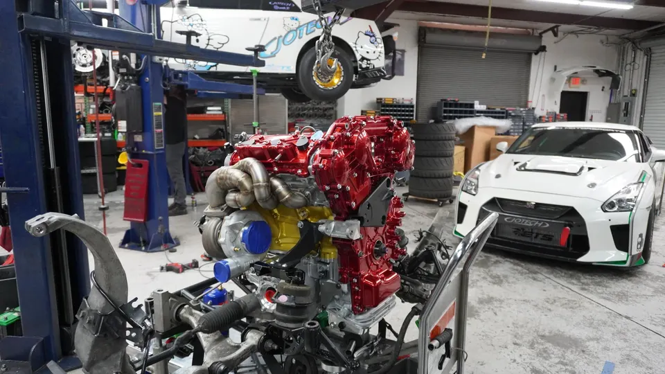 A custom built red and gold engine with a large turbocharger on a stand in a Jotech Motorsports shop, with a white Nissan GT-R on a lift and another GT-R on the
