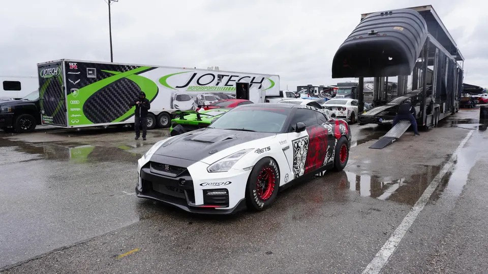 A custom white and red Nissan GT-R race car with red wheels parked on wet asphalt at a track event with Jotech Motorsports trailers.