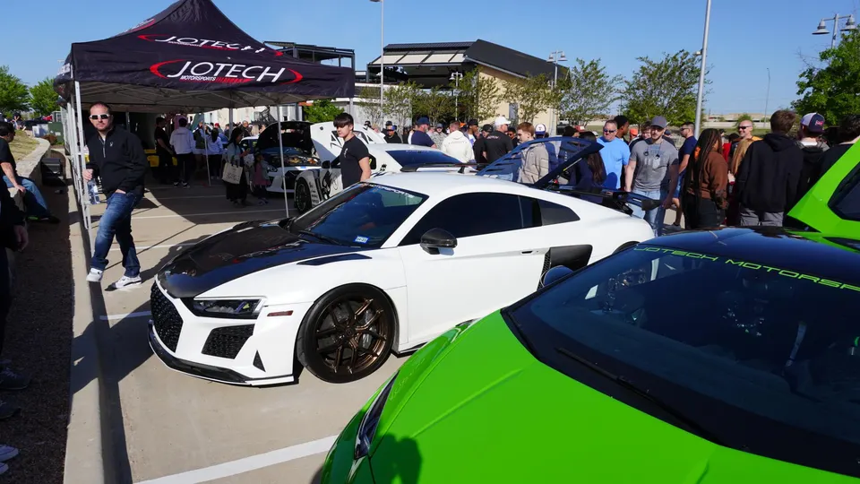 A crowd gathers around a white Audi R8 and a green Lamborghini at a Jotech Motorsports car meet.