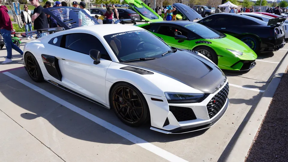 White Audi R8, green Lamborghini Huracan, and black Corvette parked at an outdoor car show with people around.