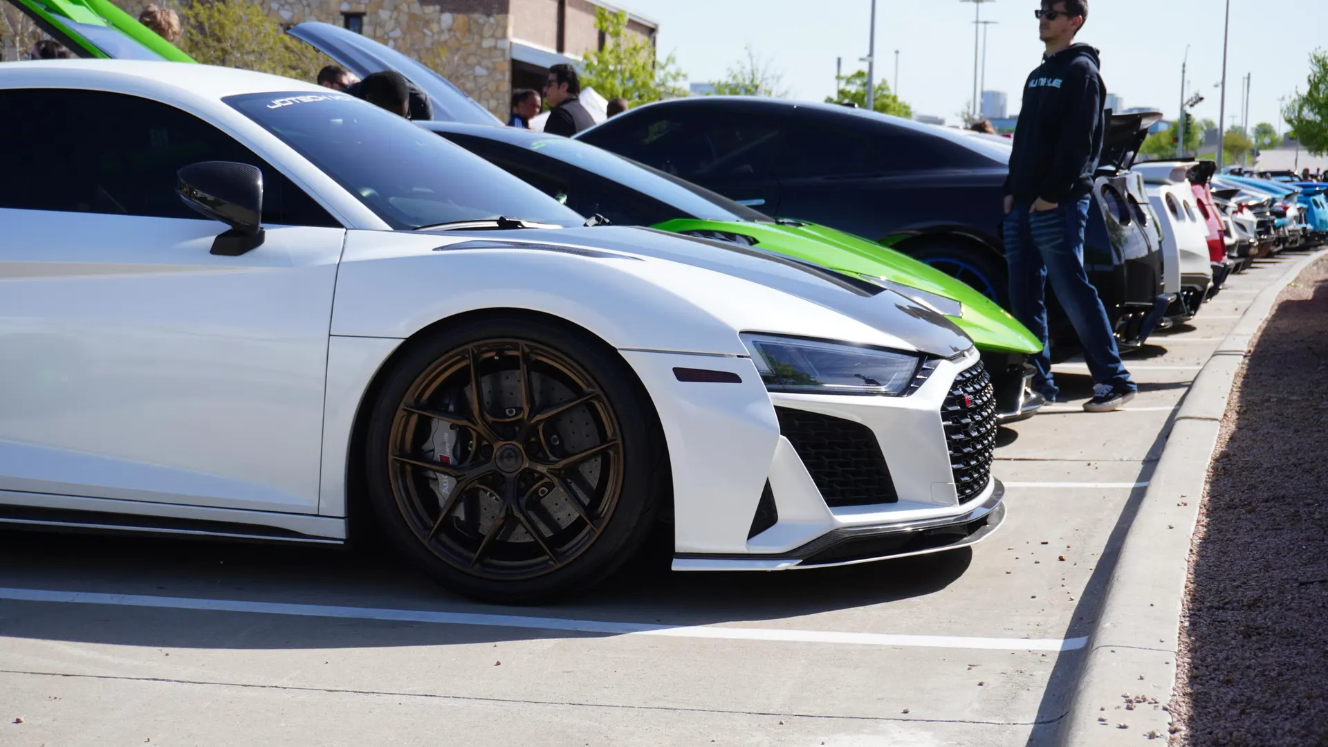 A line of high-performance cars, including a white Audi R8 and a green Lamborghini, parked at an outdoor car meet.