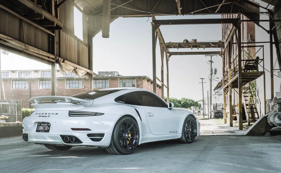 A white Porsche 911 Turbo S with black wheels parked in an industrial setting with a large metal structure overhead.