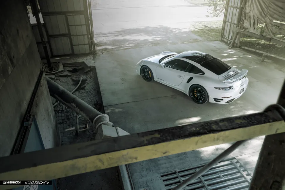 White Porsche 911 Turbo S with black aftermarket wheels and a rear wing parked in an industrial setting.