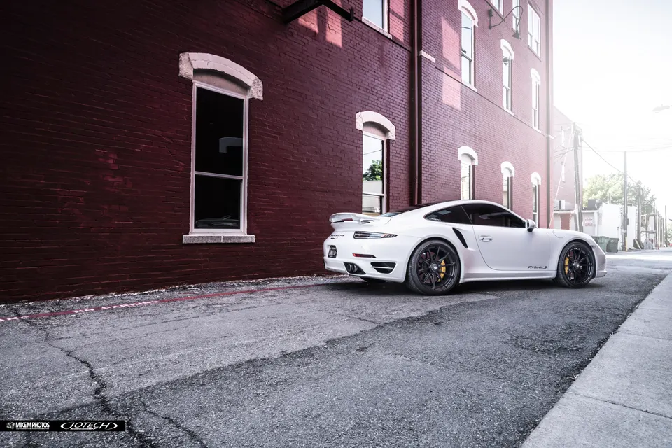 White Porsche 911 Turbo S with aftermarket wheels parked on a street next to a red brick building.
