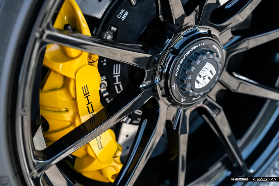 A close-up of a Porsche's black centerlock wheel with a bright yellow brake caliper and Porsche branding.