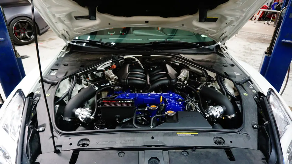Nissan GT-R engine bay with custom blue intake manifold, Boost Logic air intakes, and blow-off valves in a performance shop.