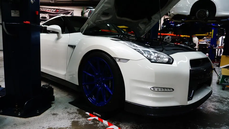 A white Nissan GT-R with blue wheels on a lift in a performance tuning shop with its hood open, ready for work.